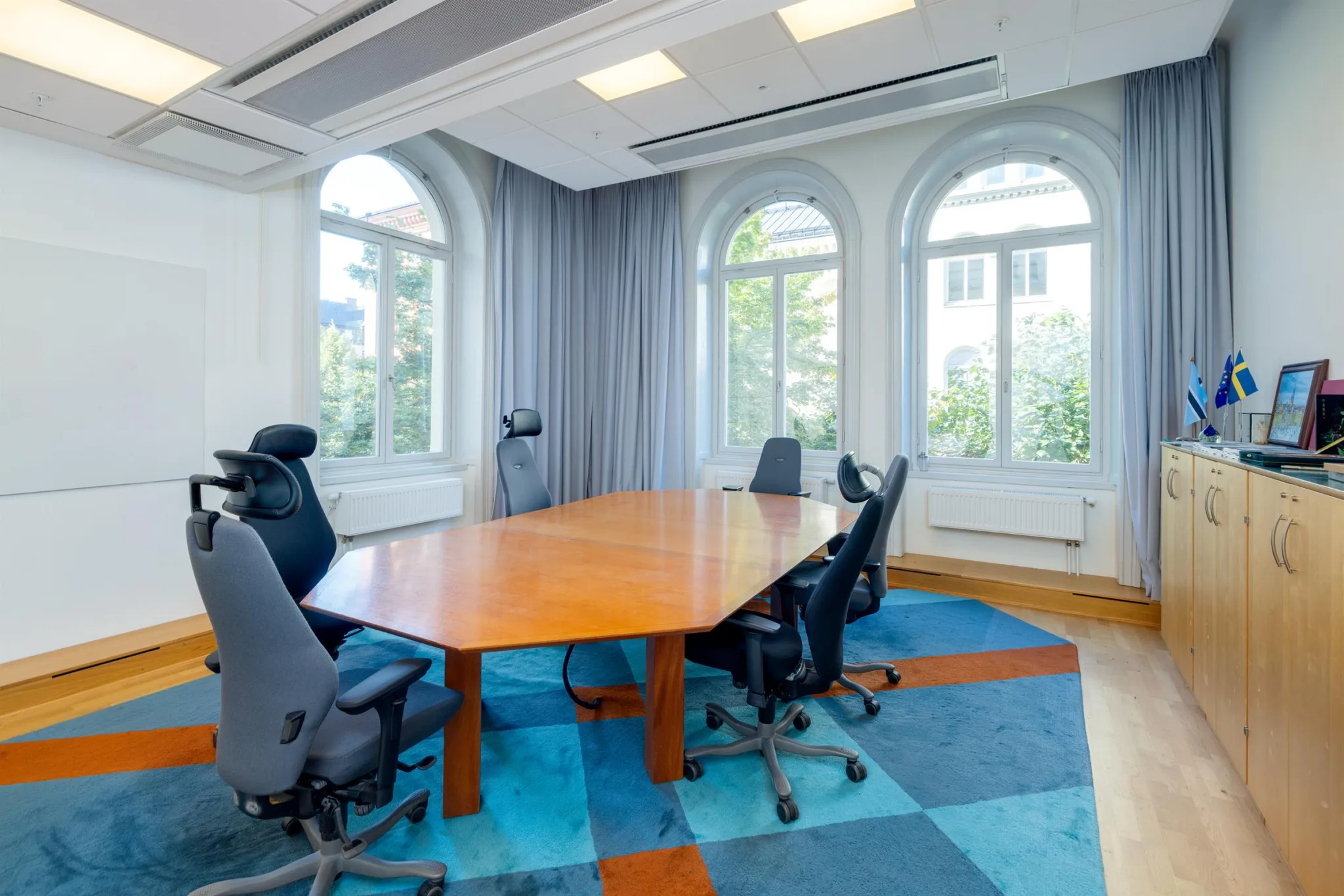Ett konferensrum med ett stort träbord, kontorsstolar och fönster med gardiner som släpper in ljus.