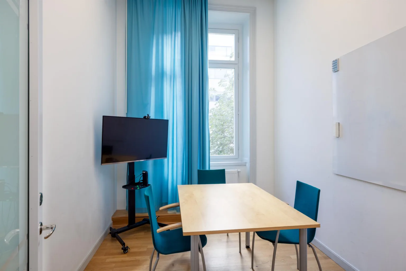 Ett litet mötesrum med ett bord, fyra stolar, en TV-skärm på stativ och blå gardiner.