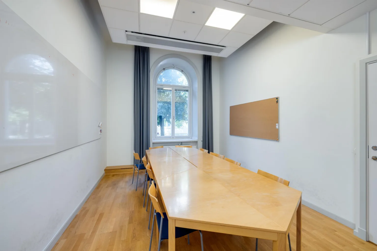 Konferensrum med långt träbord, stolar, anslagstavla och stort fönster med gardiner som släpper in naturligt ljus.