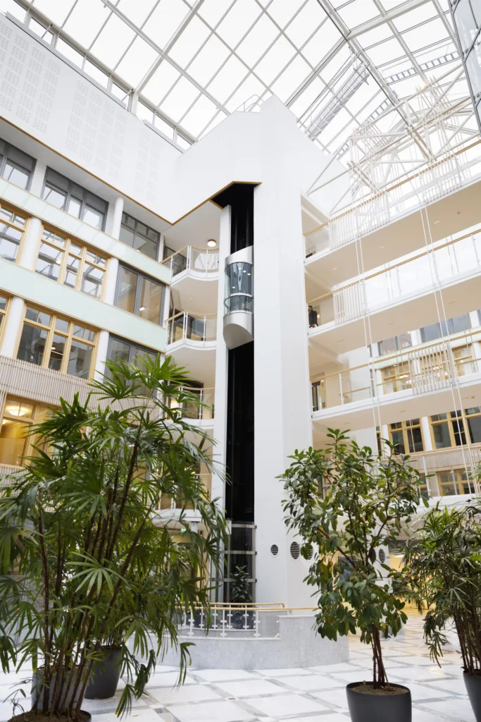 En ljus och luftig inomhusgård med glasväggar, grönska och en hiss i centrum.