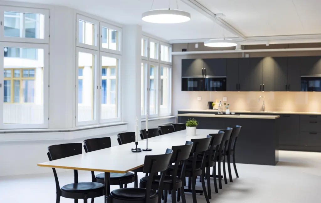 Ett modernt konferensrum med lång vit bordsskiva, svarta stolar och ett kök i bakgrunden.