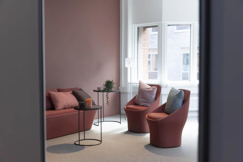 Ett stilrent loungeområde med rosa fåtöljer, en soffa, små bord och stora fönster som släpper in naturligt ljus.