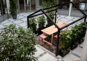 5. Ett uterum med ett träbord och rosa stolar under en svart metallstruktur, omgiven av grönskande växter.