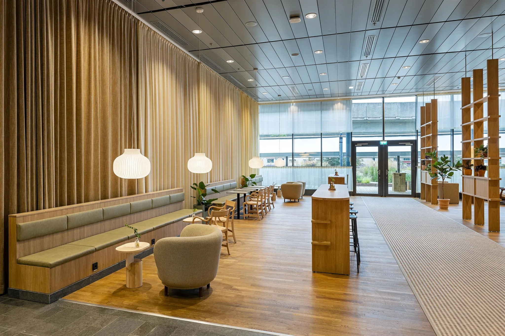 En rymlig lounge med trägolv, sittgrupper och runda lampor, stora fönster som släpper in naturligt ljus.