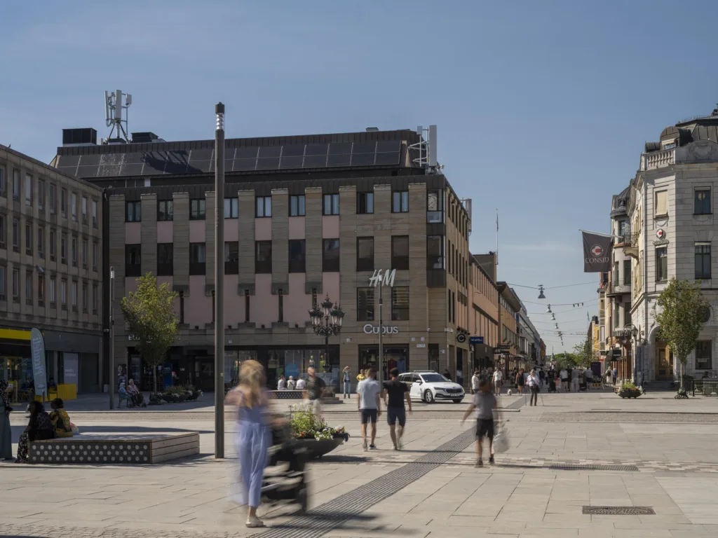 En stadsgata med människor som promenerar, omgivna av byggnader under en klarblå himmel.