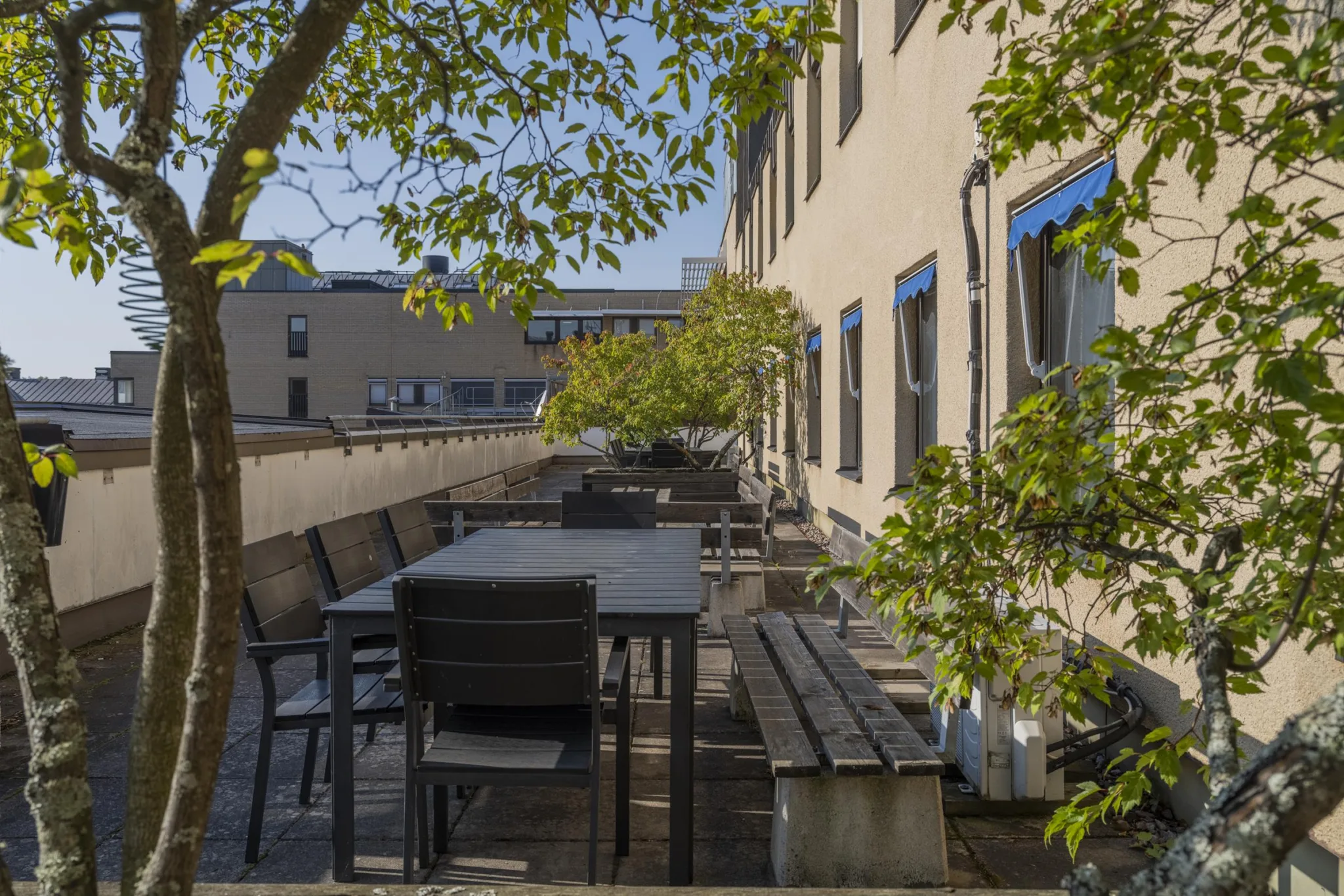 En lång terrass med flera sittplatser, bord och gröna träd mot en byggnad.