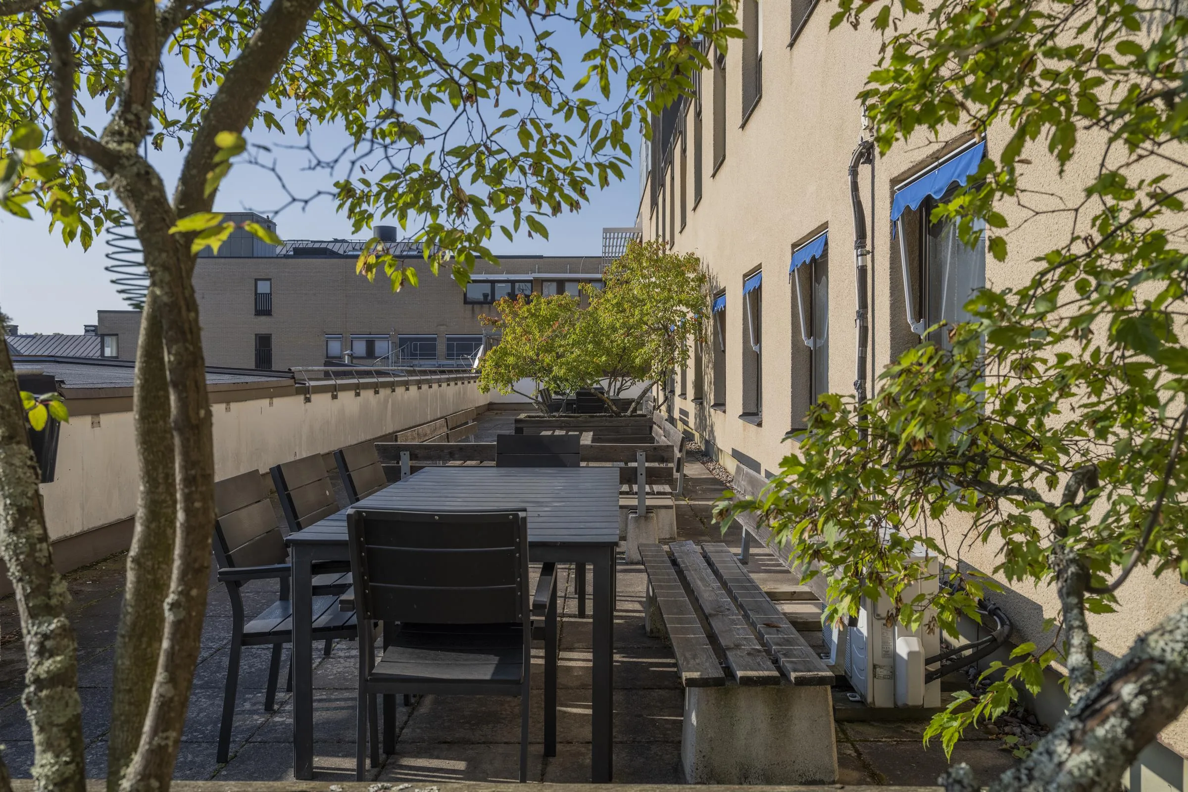 En lång terrass med flera sittplatser, bord och gröna träd mot en byggnad.