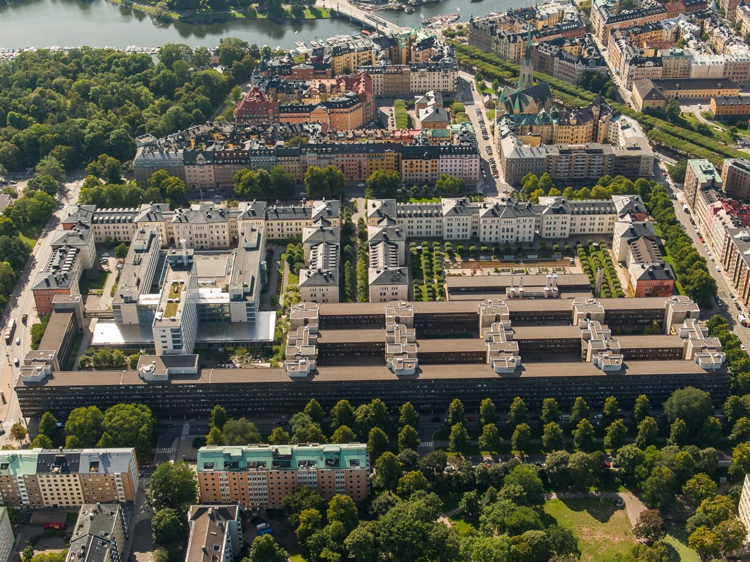 Flygfoto över stadsdel med symmetriskt placerade byggnader och grönområden, en flod syns i bakgrunden.