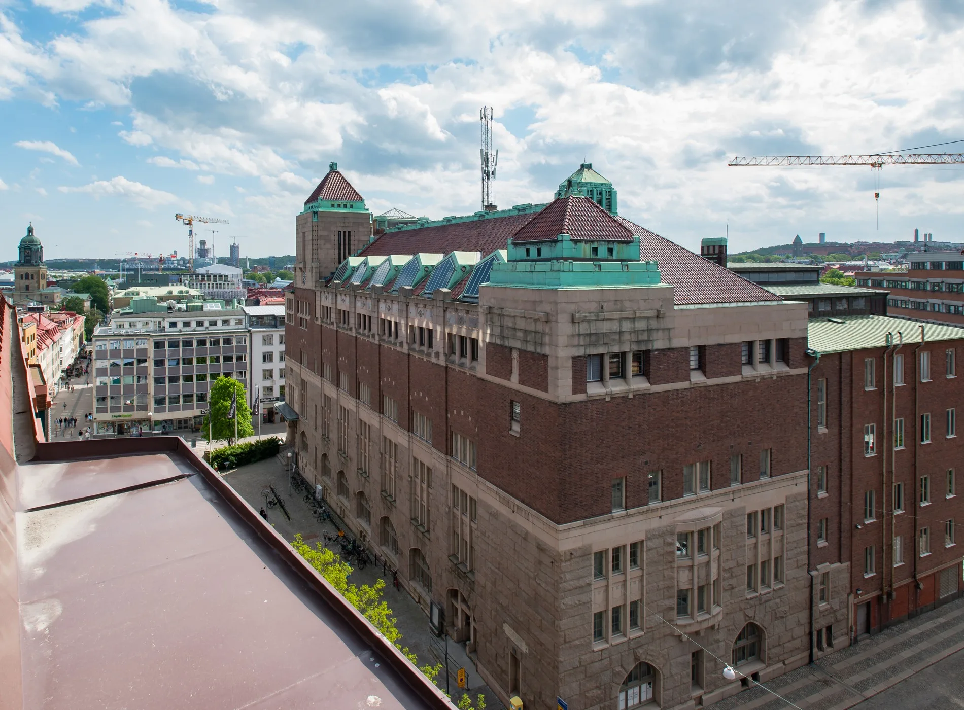 4. En stor byggnad i rödbrunt tegel med grönt tak och torn, omgiven av andra byggnader och en molnig himmel i bakgrunden.