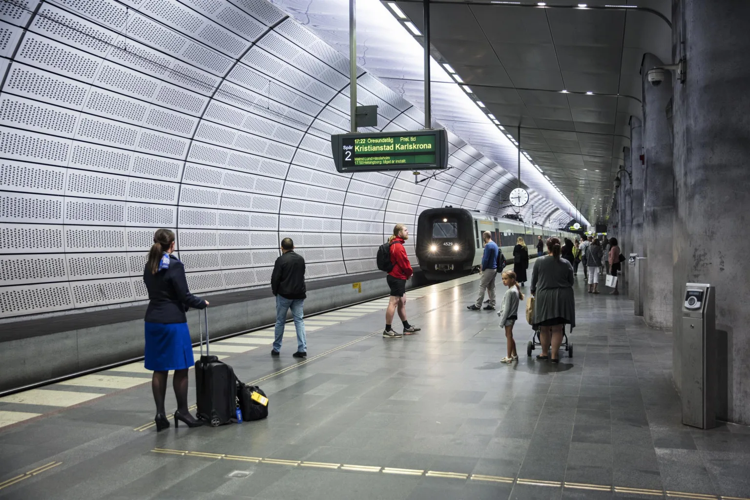 En tunnelbanestation med människor som väntar på perrongen och ett tåg som närmar sig.