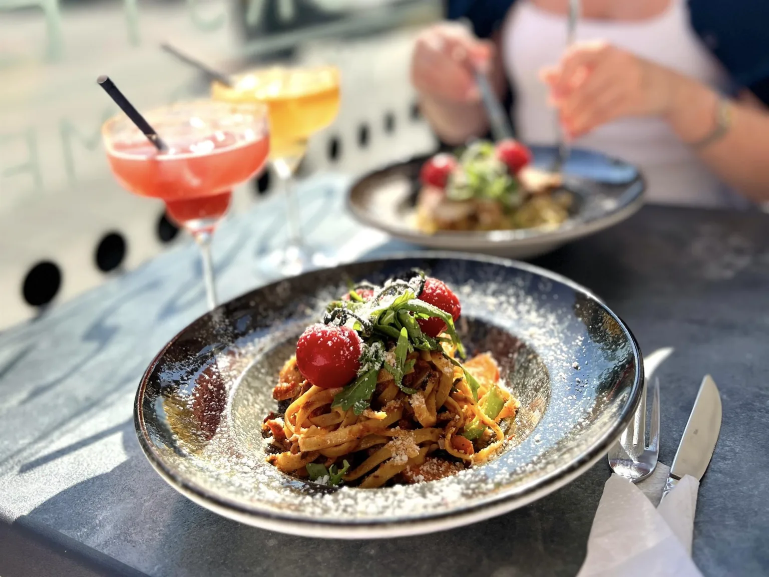 Två tallrikar med pasta och körsbärstomater, samt cocktails på ett bord utomhus.