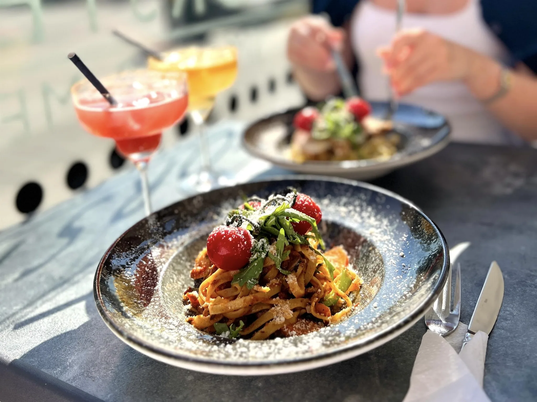 Två tallrikar med pasta och körsbärstomater, samt cocktails på ett bord utomhus.
