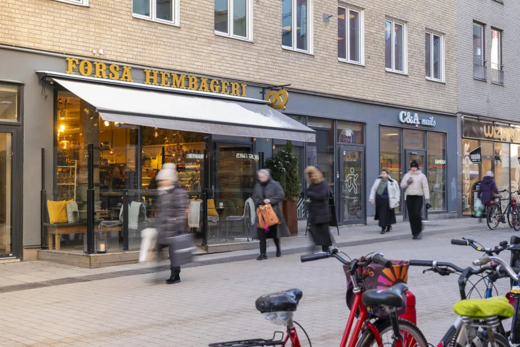 En stadsgata med människor som passerar förbi butiker, däribland en med skylten "Örsa Hemkafé." En cykel står parkerad i förgrunden.
