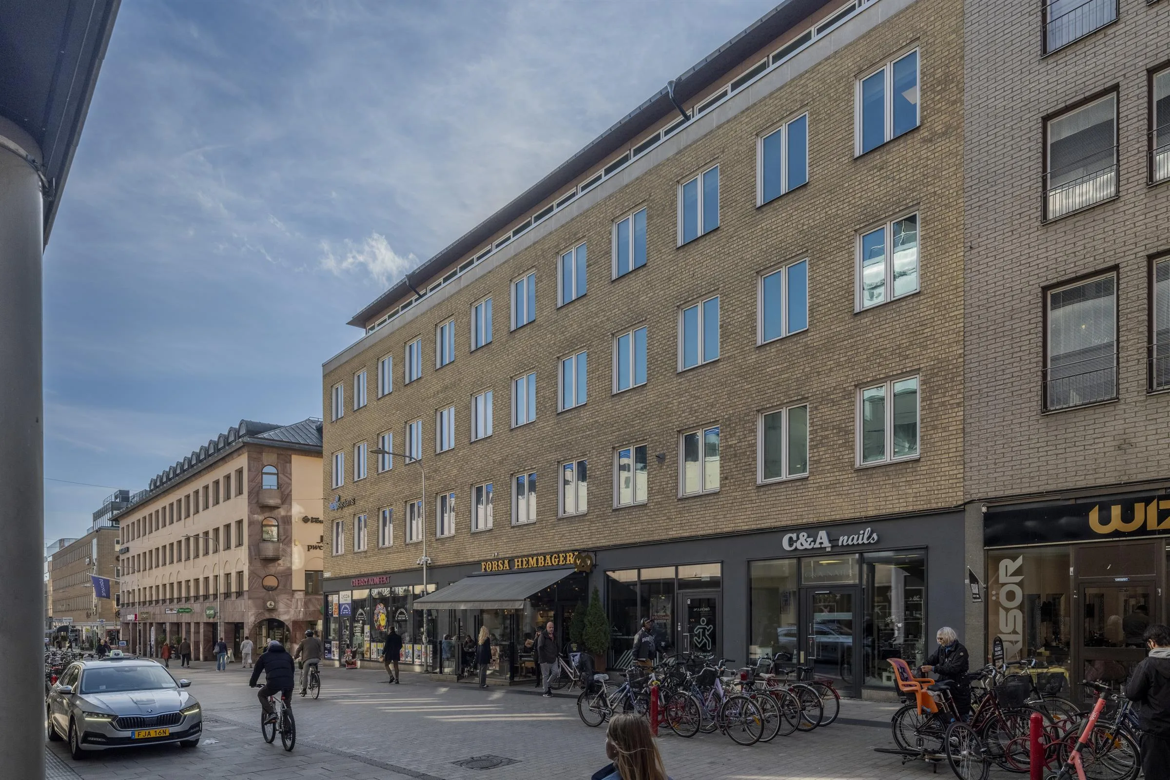 En flerbostadsbyggnad med tegelväggar och stora fönster, belägen vid en stadsgata med fotgängare och cyklar.