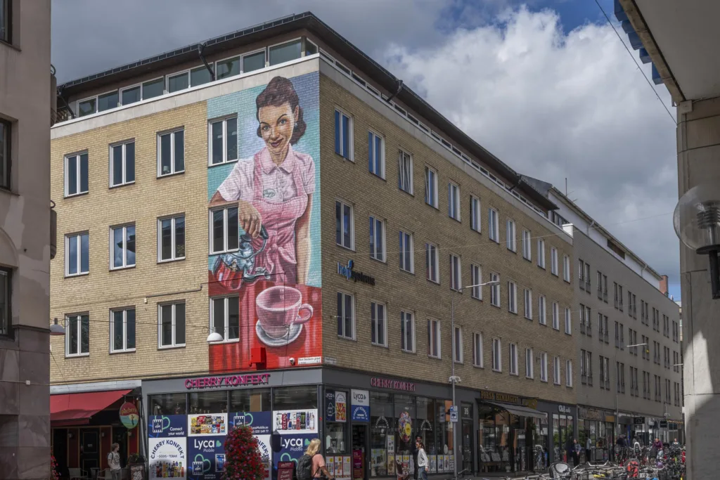 Byggnad med stor väggmålning av en kvinna som brygger kaffe, omgiven av butiker och skyltar under en molnig himmel.