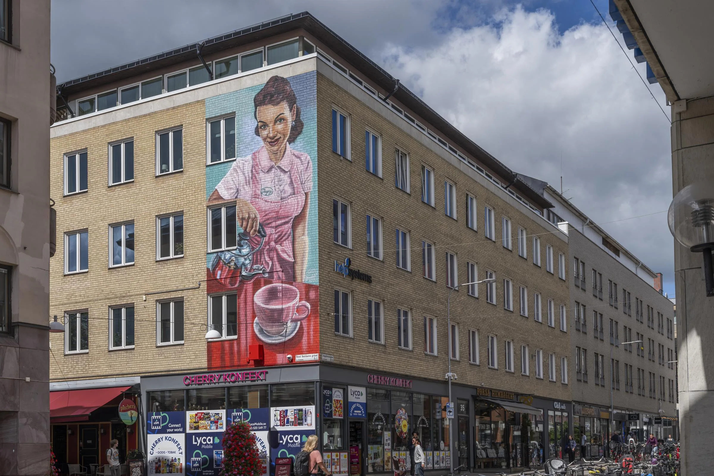 Byggnad med stor väggmålning av en kvinna som brygger kaffe, omgiven av butiker och skyltar under en molnig himmel.