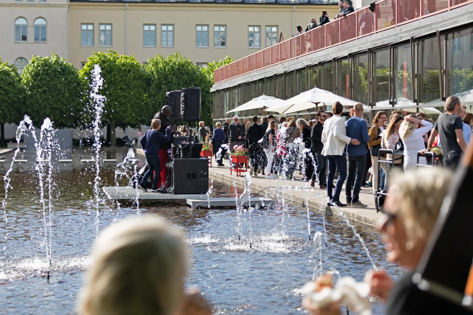 1. En utomhusmiljö med fontäner i förgrunden, människor som umgås vid bord och en musiker som spelar vid ett torg.