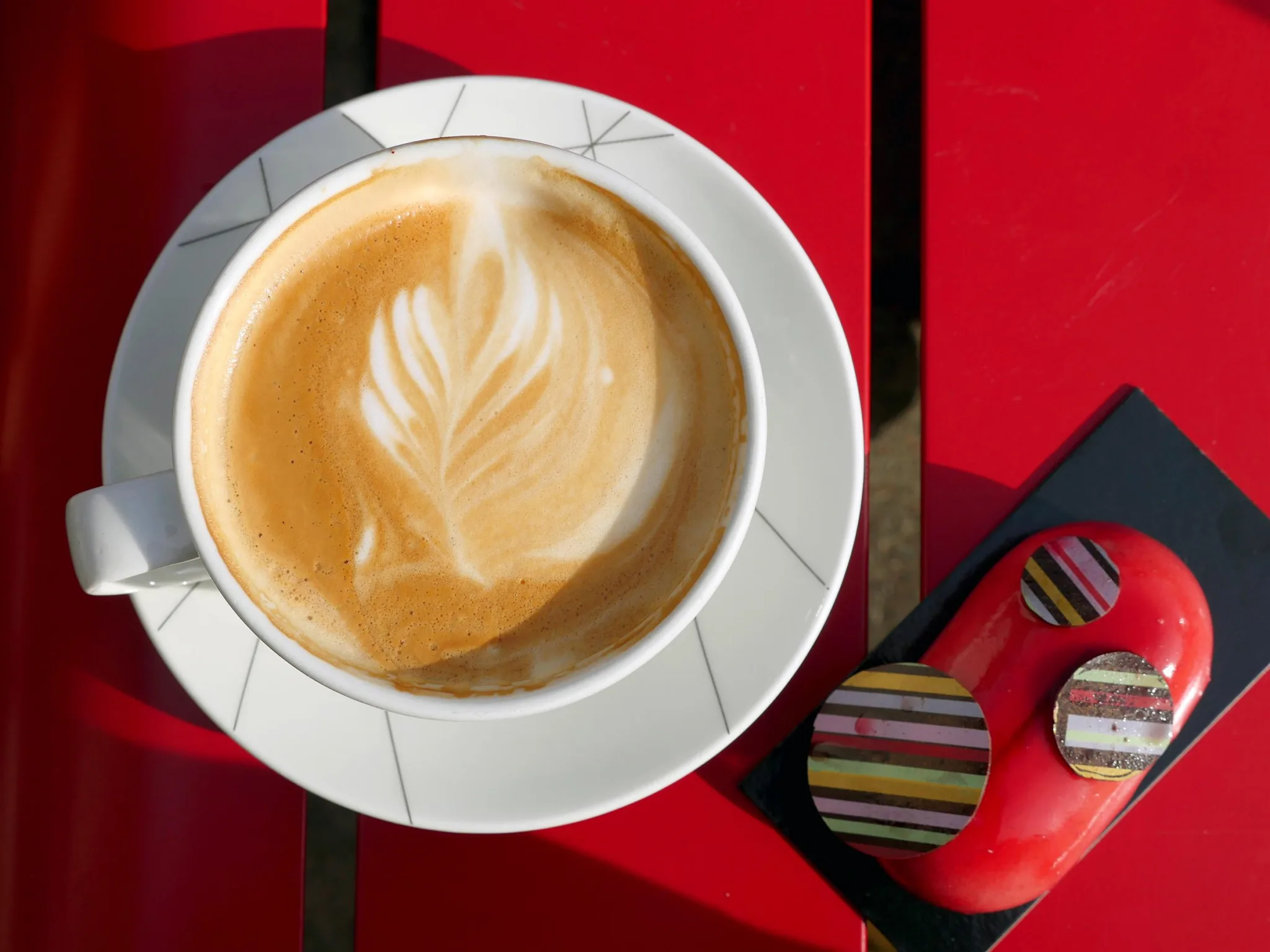 3. En kopp cappuccino med latte art i form av ett blad, stående på ett rött bord bredvid en randig godisförpackning.