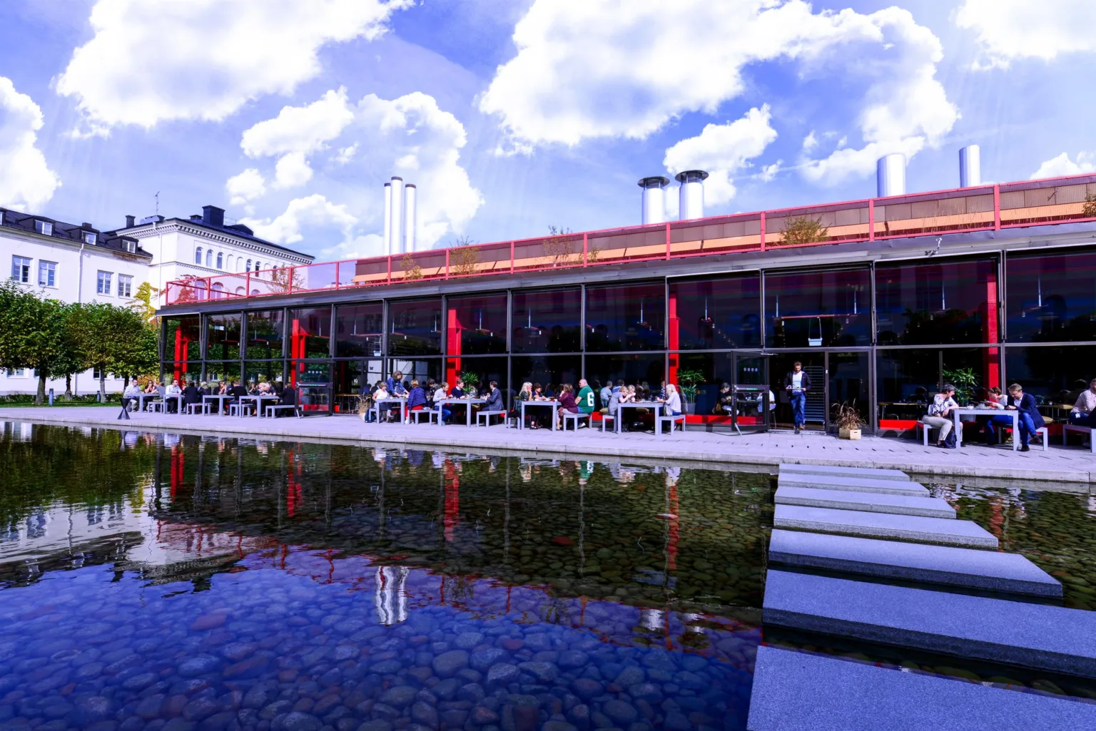 En modern byggnad med stora fönster, röd dekoration och en reflekterande vattenyta framför.