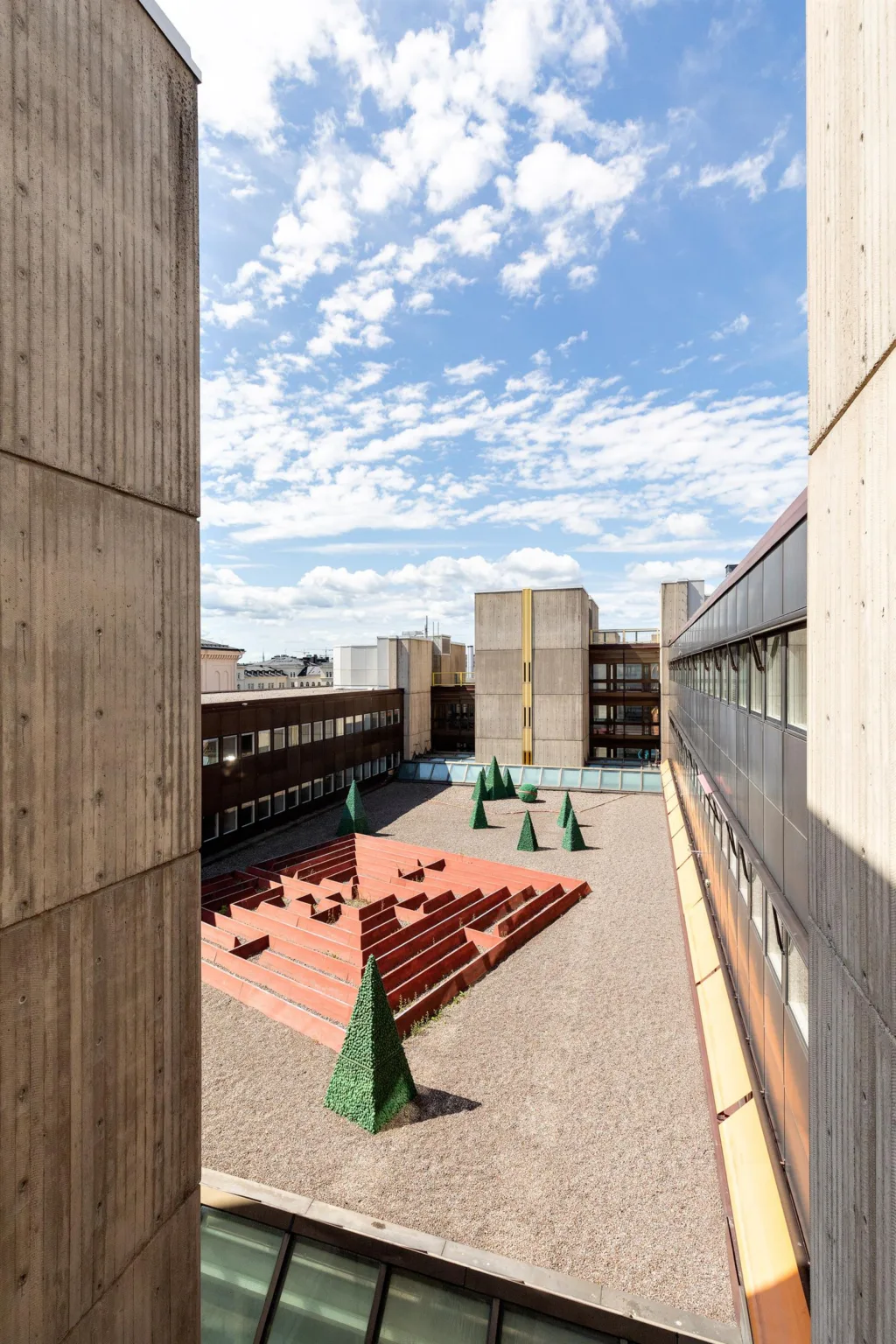 1. En innergård omgiven av betongbyggnader med sittplatser och konformade gröna träd under en klarblå himmel.