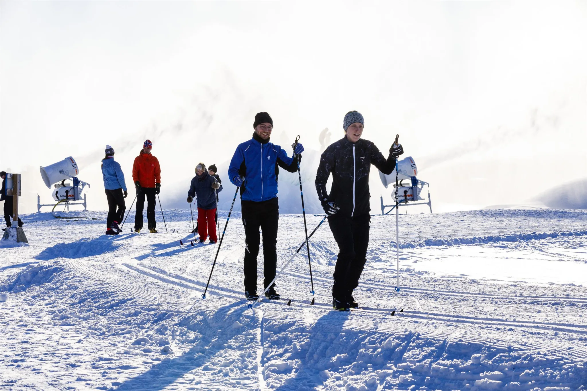 Två personer åker längdskidor på en snötäckt slätt med fler skidåkare i bakgrunden.