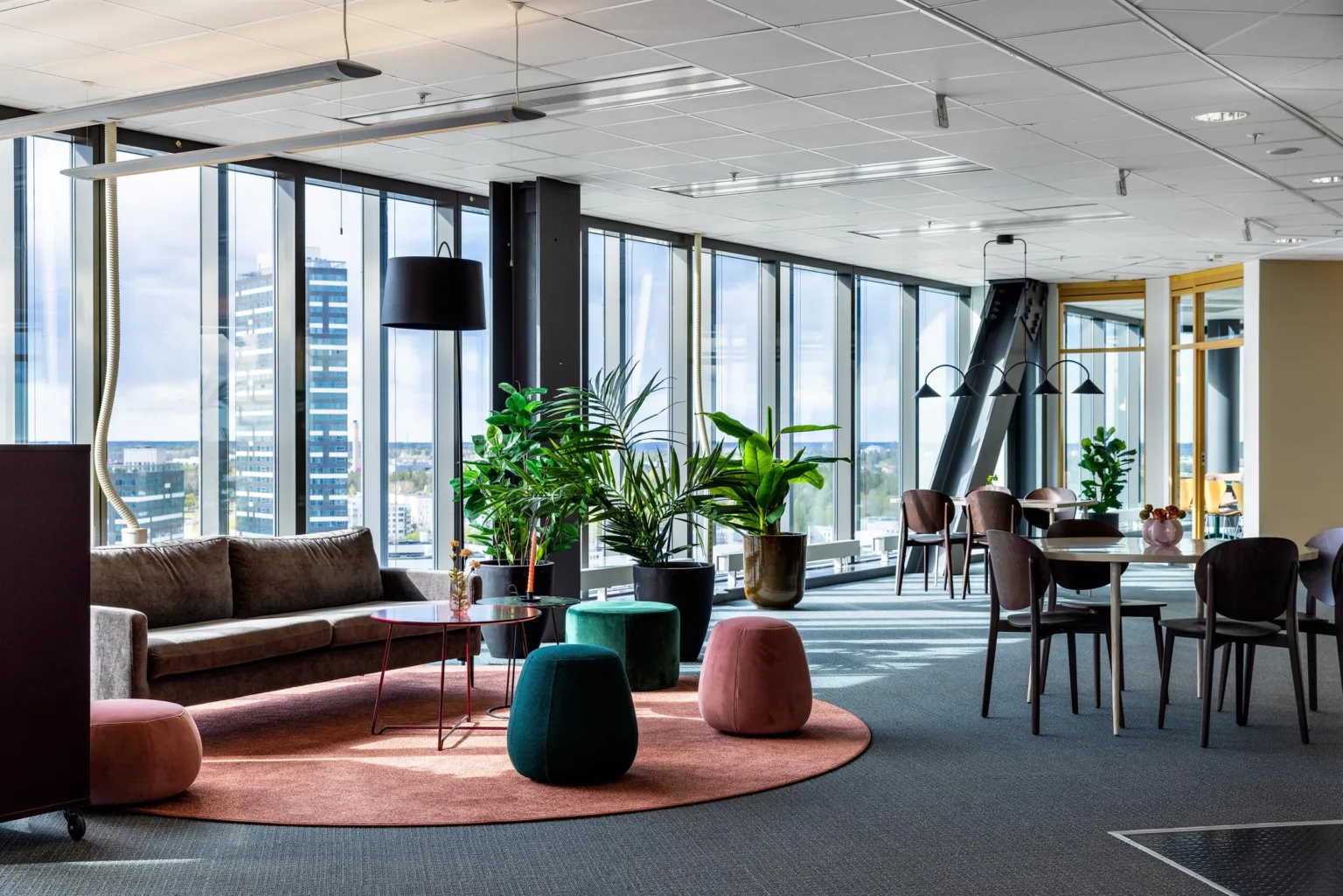 En modern lounge med soffa, sittpuffar, gröna växter och matbord nära stora glasfönster med utsikt.
