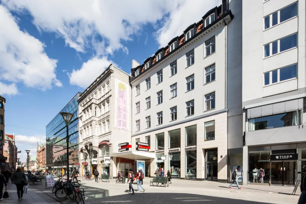 Gata med vit byggnad och angränsande butiker, personer promenerar och cyklar under blå himmel.