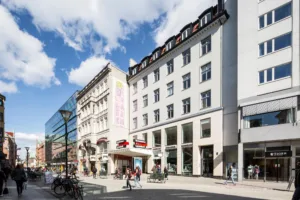 Gata med vit byggnad och angränsande butiker, personer promenerar och cyklar under blå himmel.