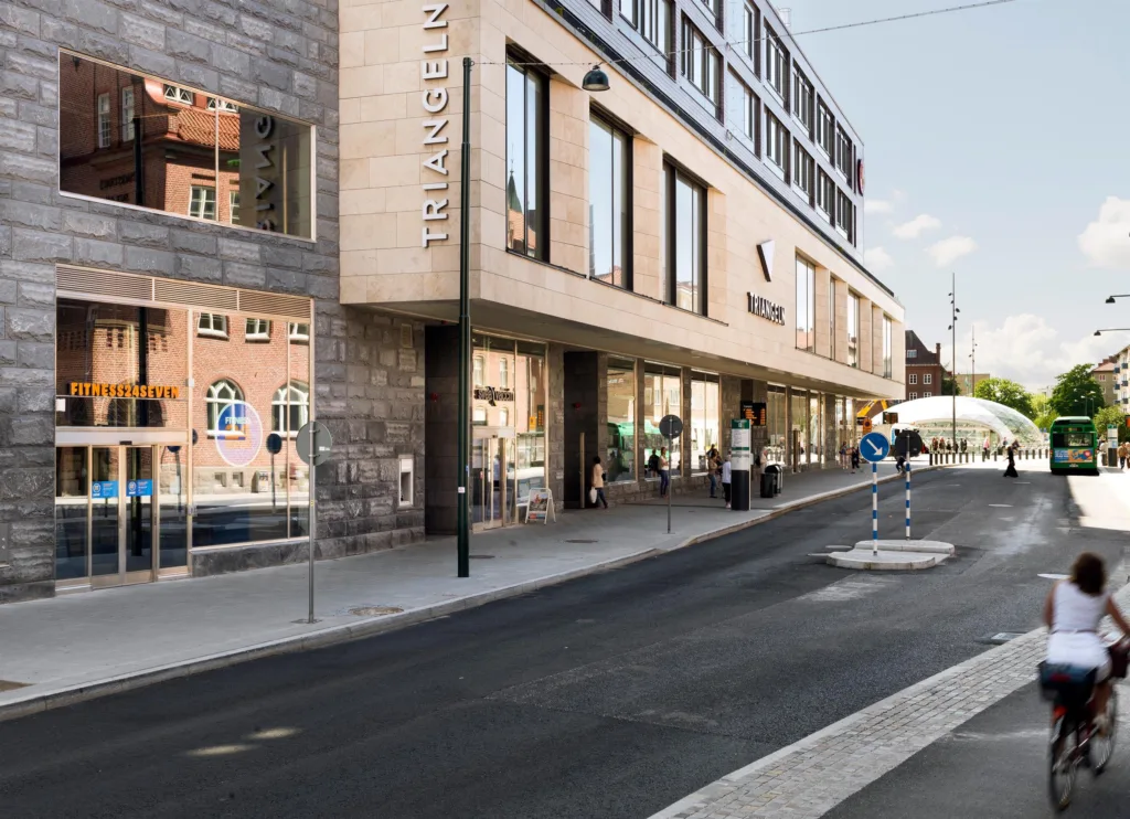 En byggnad med stora glasfönster och skylt med texten "Triangeln", omgiven av en trottoar och människor.