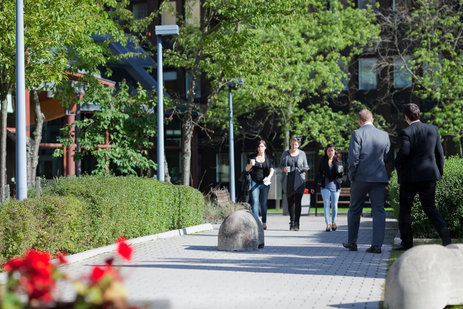 En utomhusmiljö med människor som promenerar på en gångväg omgiven av grönska och blommor.