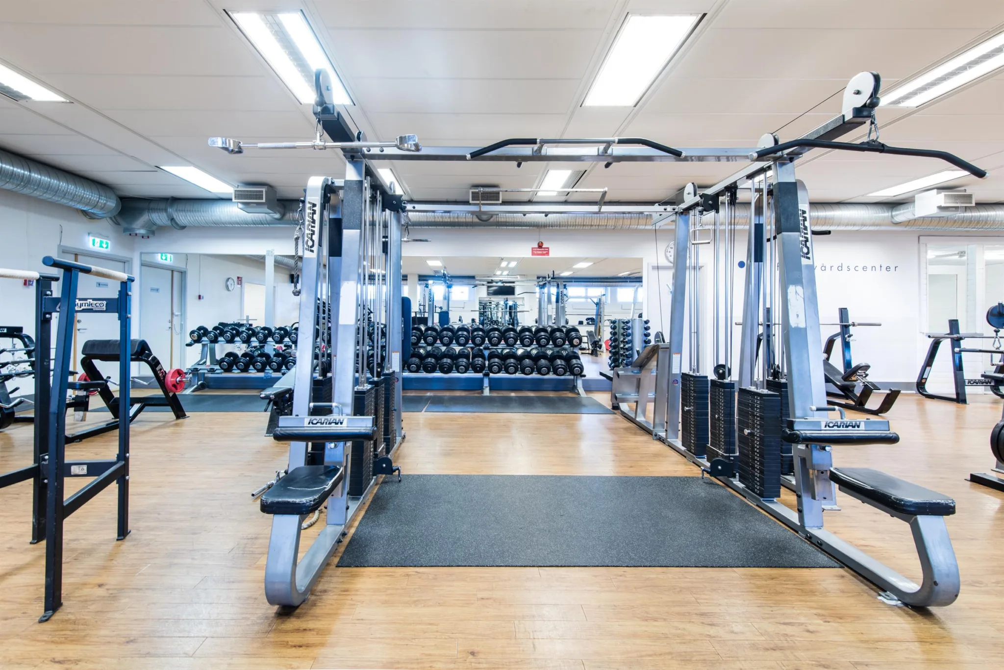 Ett gym med träningsutrustning, inklusive styrketräningsmaskiner, vikter och speglar, på ett trägolv.