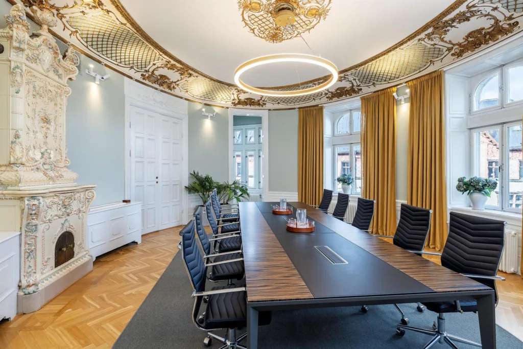 Konferensrum med lång mörk bordsskiva, svarta stolar, högt dekorerat tak och stora fönster med gardiner.