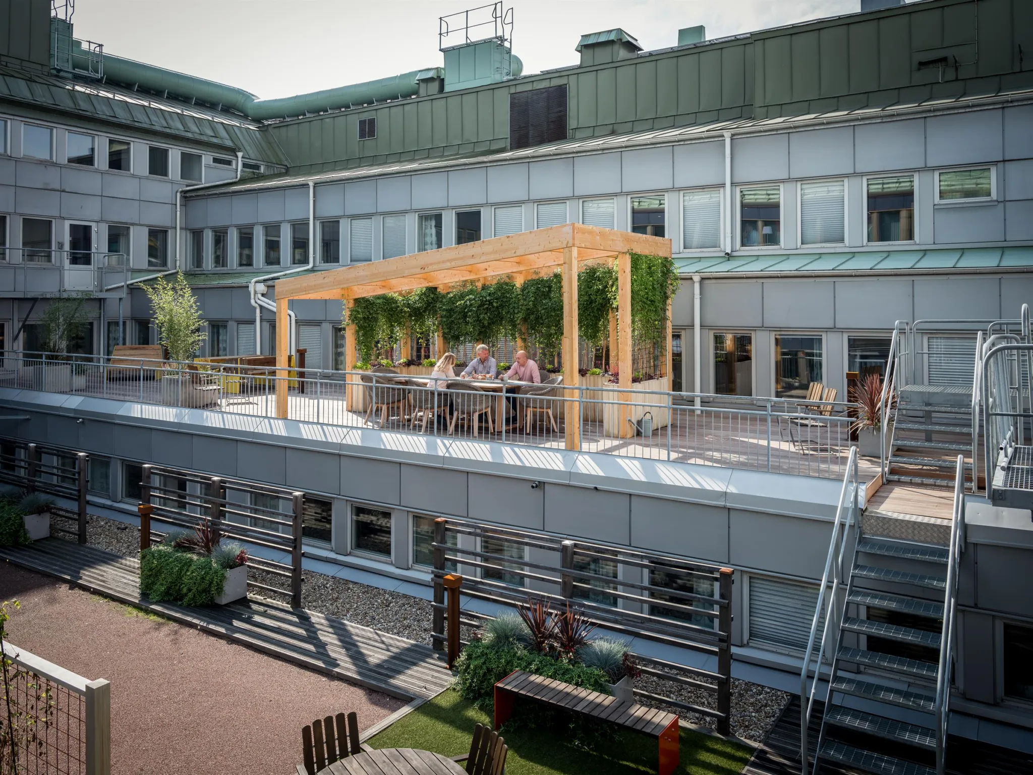 En takterrass med pergola, grönskande väggar och människor som sitter vid ett bord.