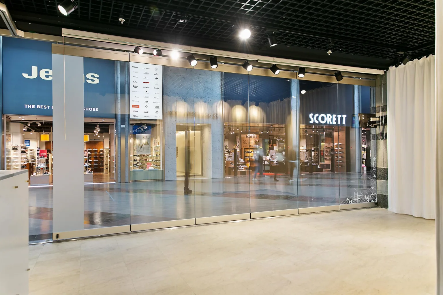 En modern butiksgalleria med glasväggar, synliga skyltar och upplysta butiker inuti.