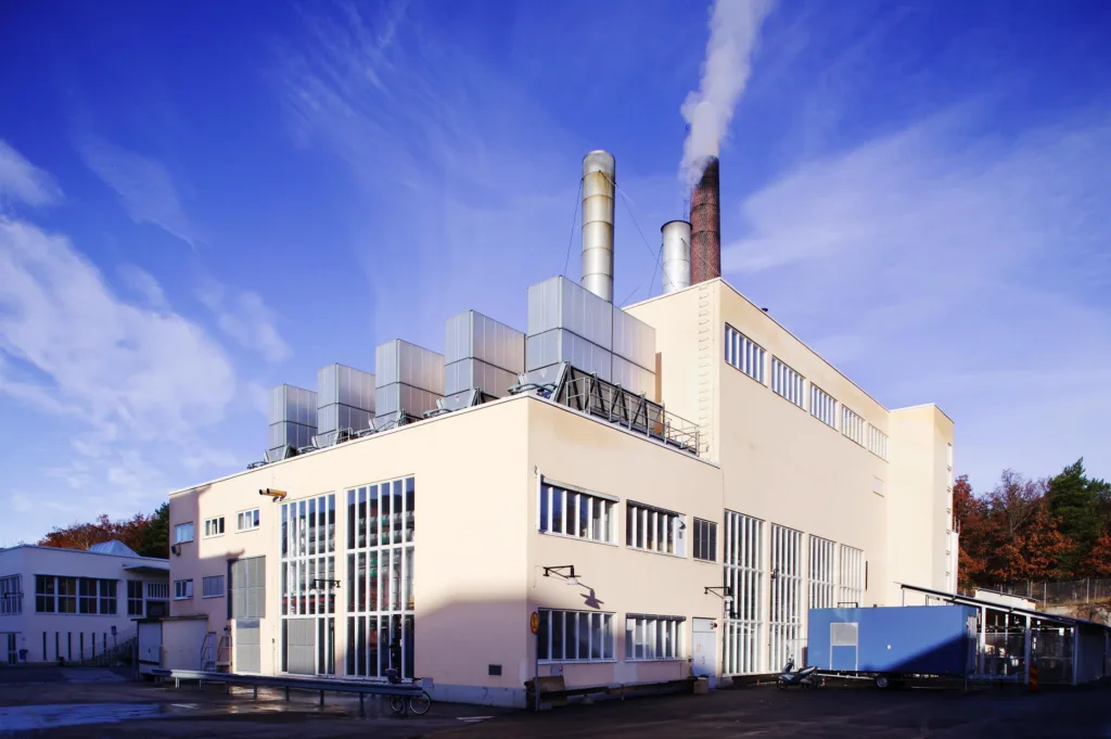 En industribyggnad med flera skorstenar som släpper ut rök, mot en blå himmel.