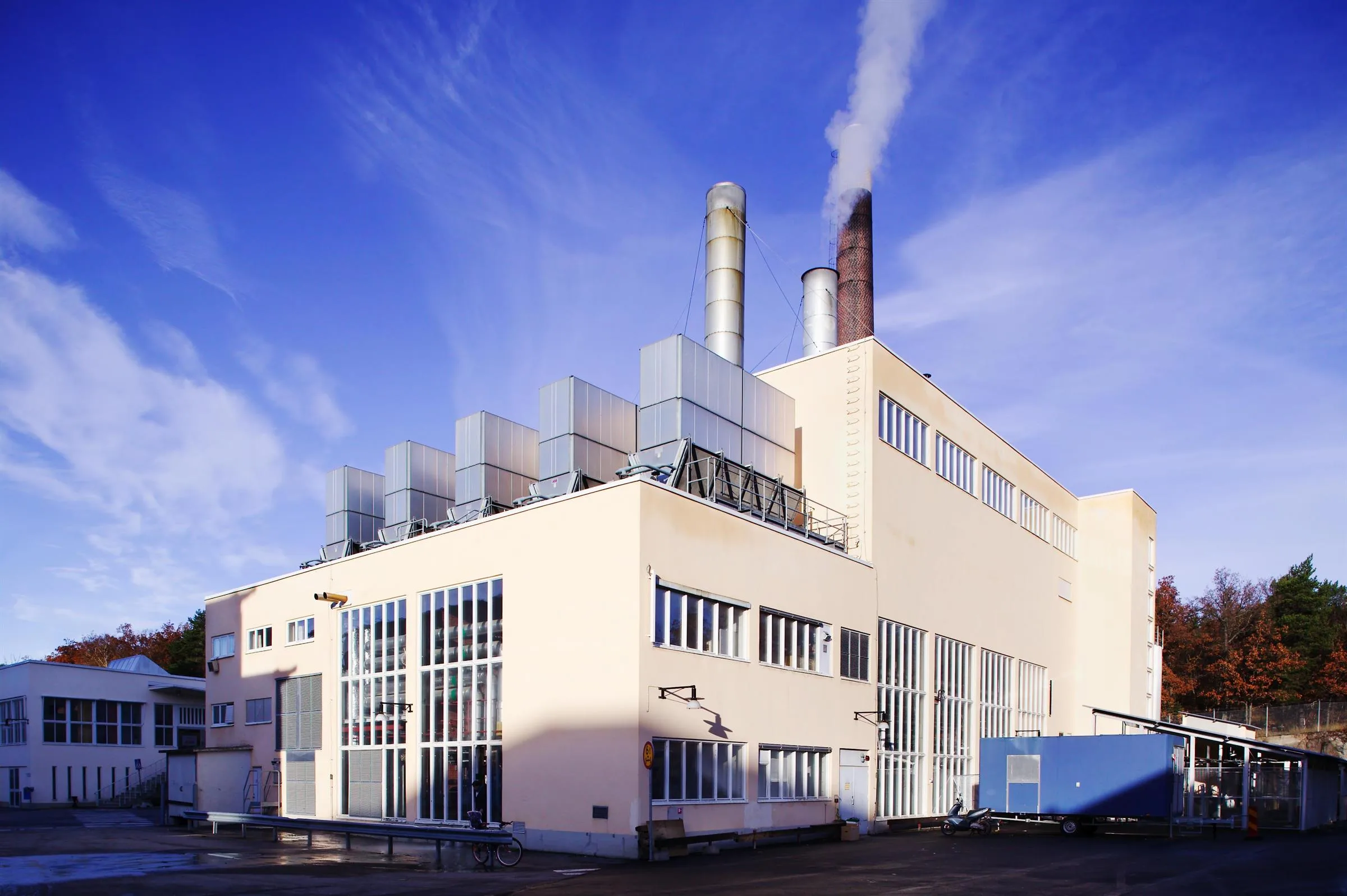 En industribyggnad med flera skorstenar som släpper ut rök, mot en blå himmel.