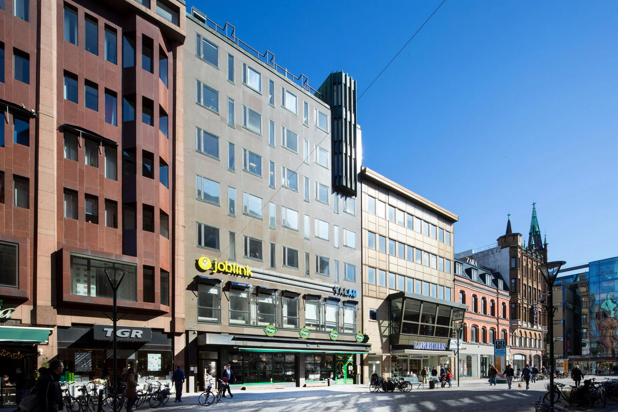 En modern byggnad med flera våningar, stora glasfönster och företagsloggor på fasaden, under en klarblå himmel.
