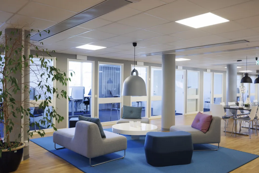 En rymlig lounge med soffor, bord, växter och en stor hängande lampa i ett kontorsutrymme.