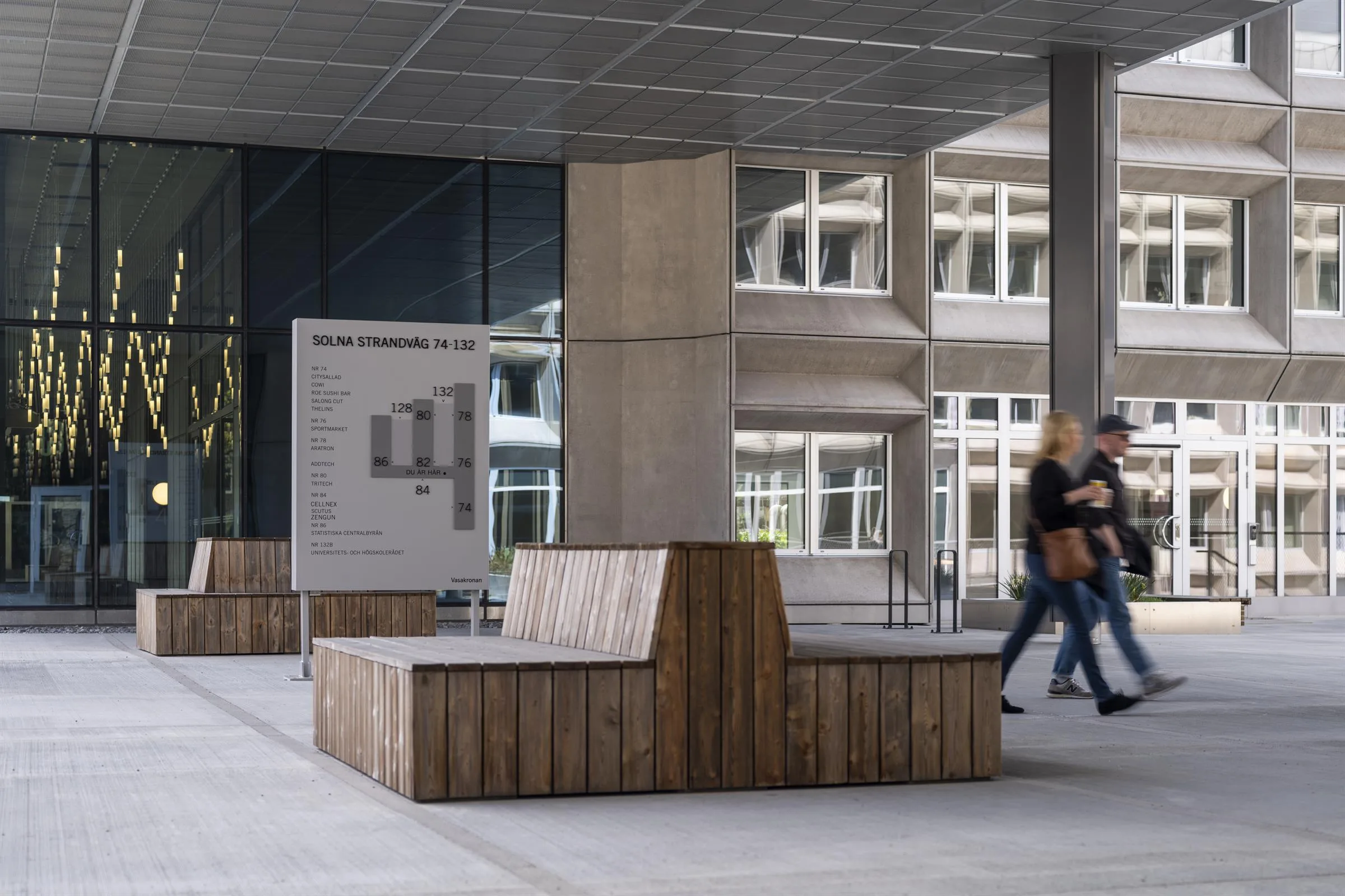 Träbänkar under en byggnads överhängande tak med en skylt i bakgrunden; två personer går förbi.