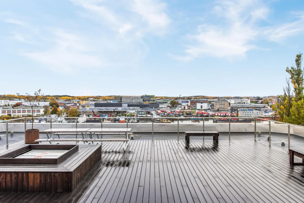 En stor takterrass med trägolv, bänkar och utsikt över en stad med byggnader och höstfärgade träd.