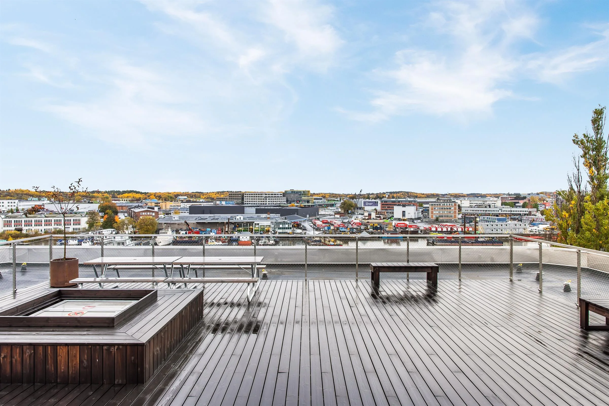En stor takterrass med trägolv, bänkar och utsikt över en stad med byggnader och höstfärgade träd.