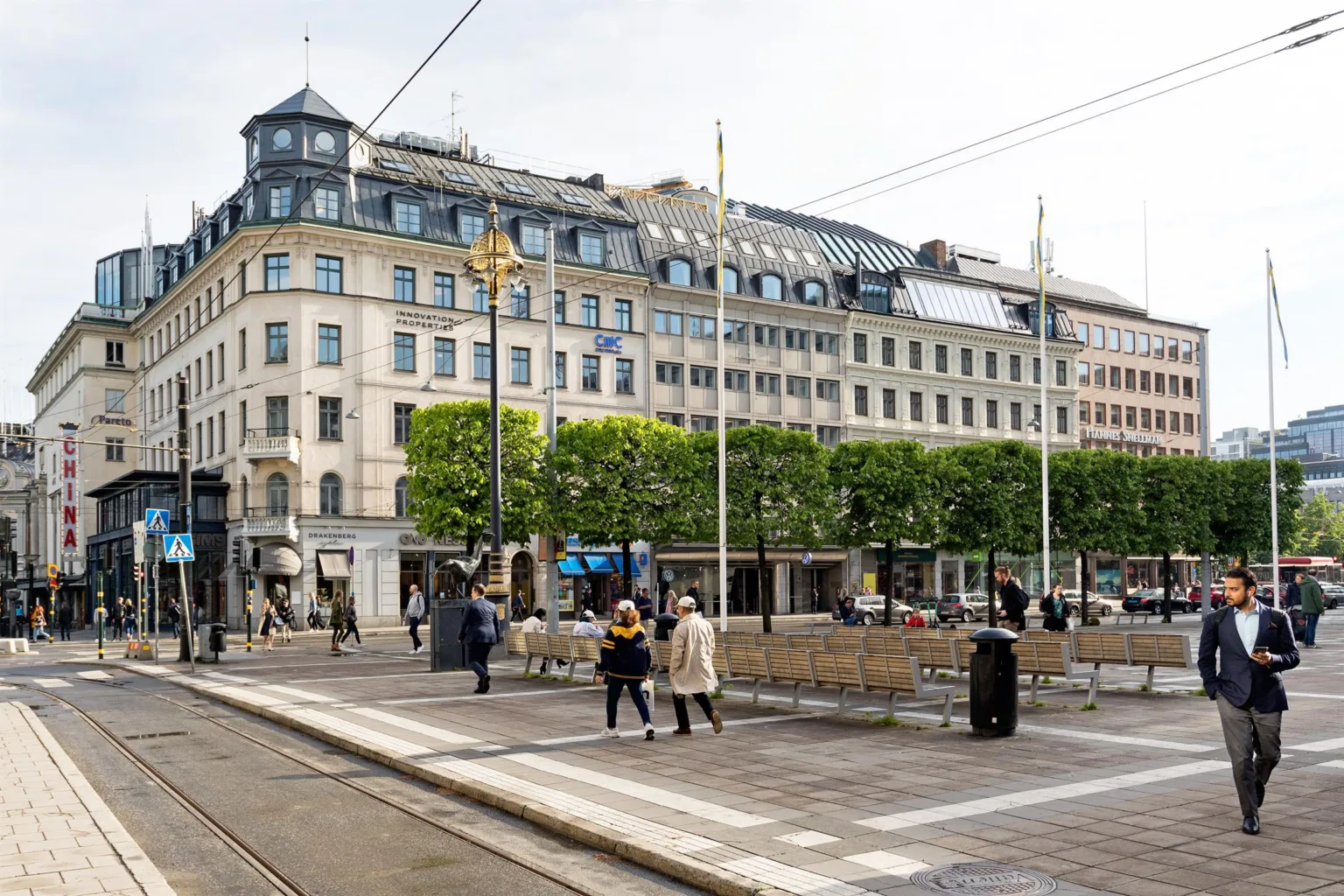 En stadsgata med människor som promenerar, spårvagnsspår och en rad byggnader med grönskande träd framför.