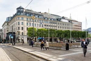 En stadsgata med människor som promenerar, spårvagnsspår och en rad byggnader med grönskande träd framför.