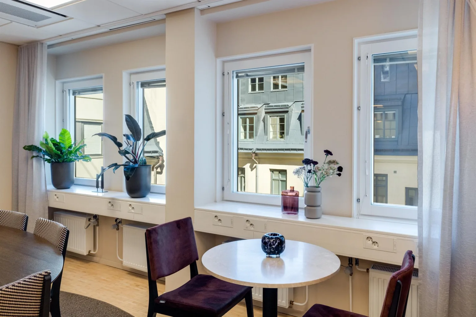 Ett litet bord med två stolar framför fönster; krukväxter och utsikt mot en byggnad utanför.