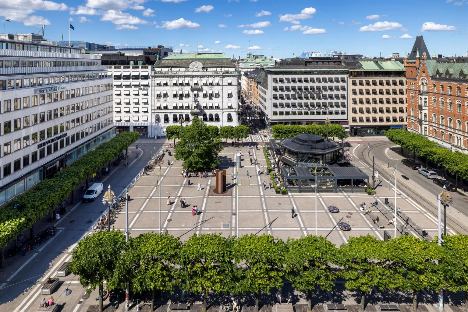 En öppen plats i stadsmiljö omgiven av byggnader med träd och bänkar, samt människor som rör sig över torget under en klarblå himmel.