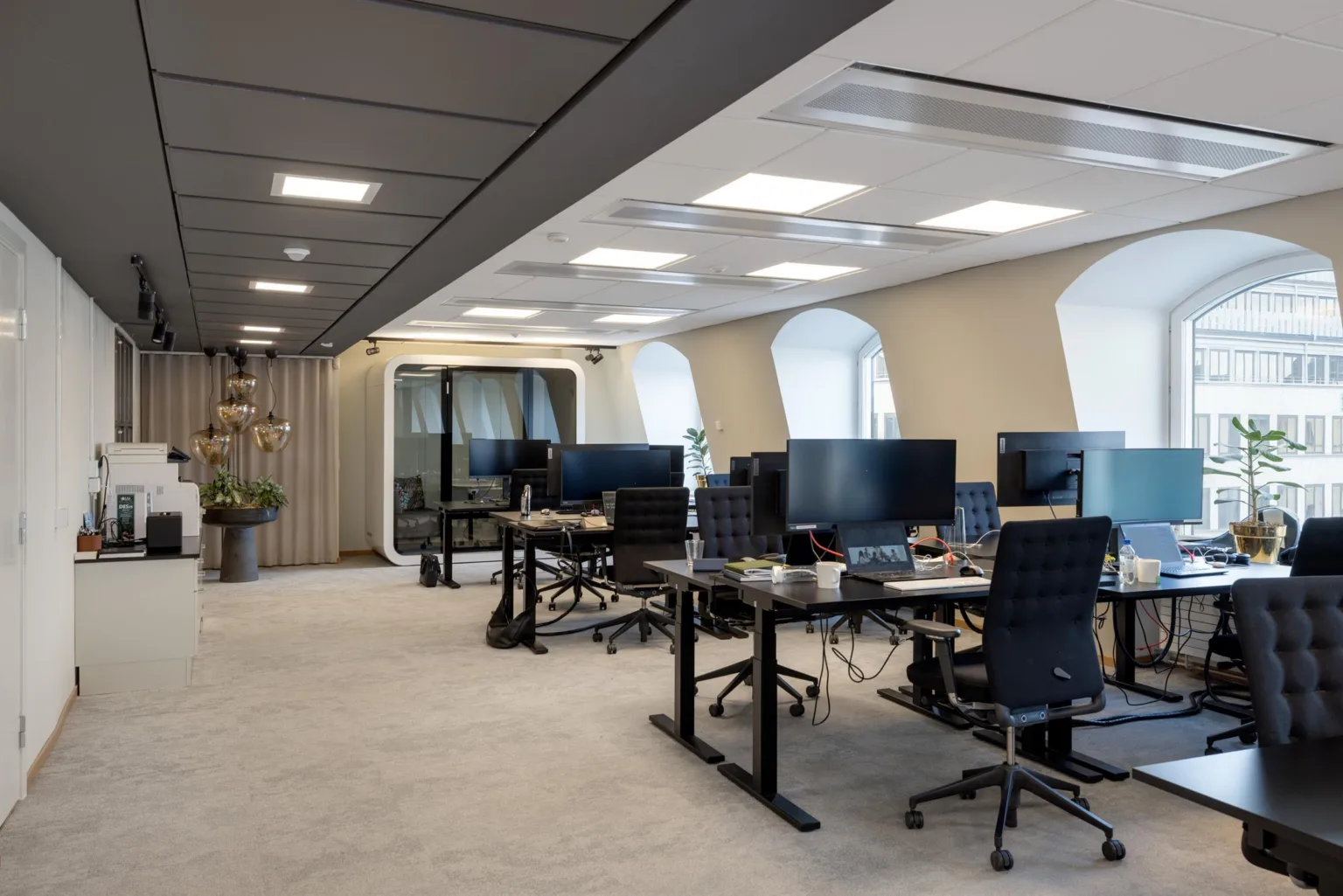 Ett modernt kontorsutrymme med skrivbord, datorskärmar och stolar i ett ljust rum med stora fönster och ljudisolerade mötesbås.