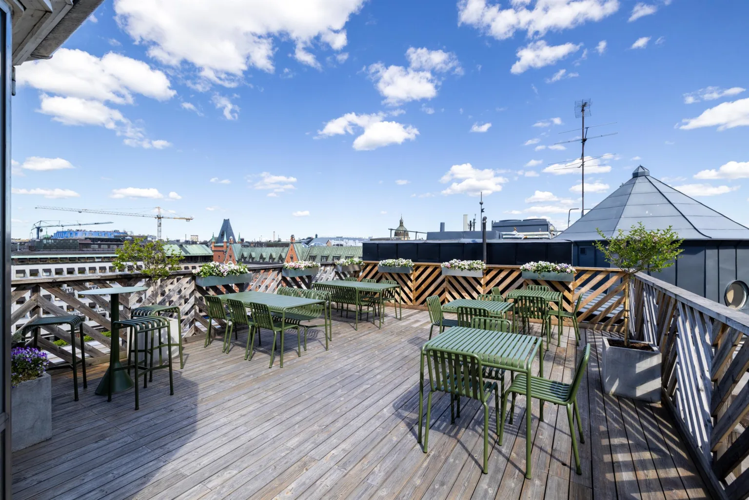 En takterrass med bord och stolar, trägolv och utsikt över stadens byggnader under en klarblå himmel med vita moln.