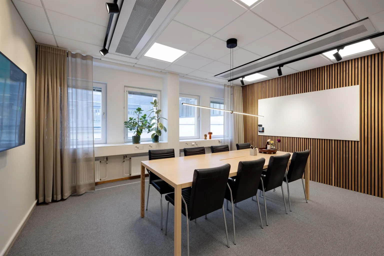 Ett konferensrum med ett långt bord, åtta stolar, en whiteboard, stora fönster och gardiner.