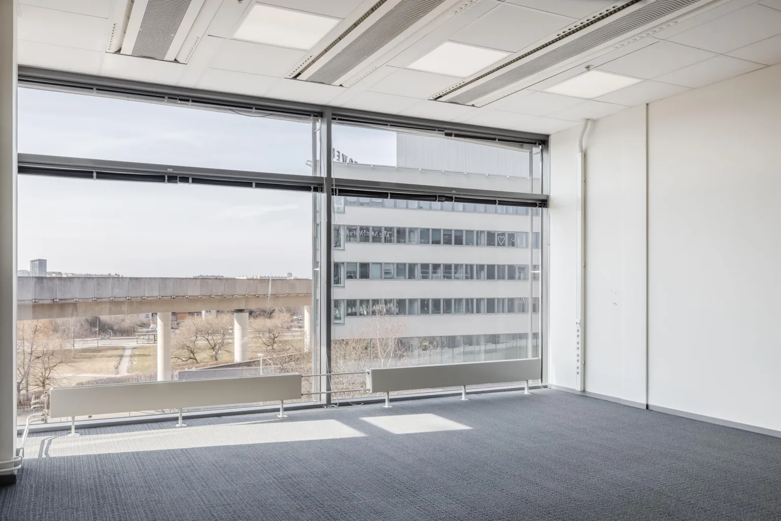 Ett kontorsrum med stora fönster som släpper in naturligt ljus och en utsikt över ett kontorskomplex.