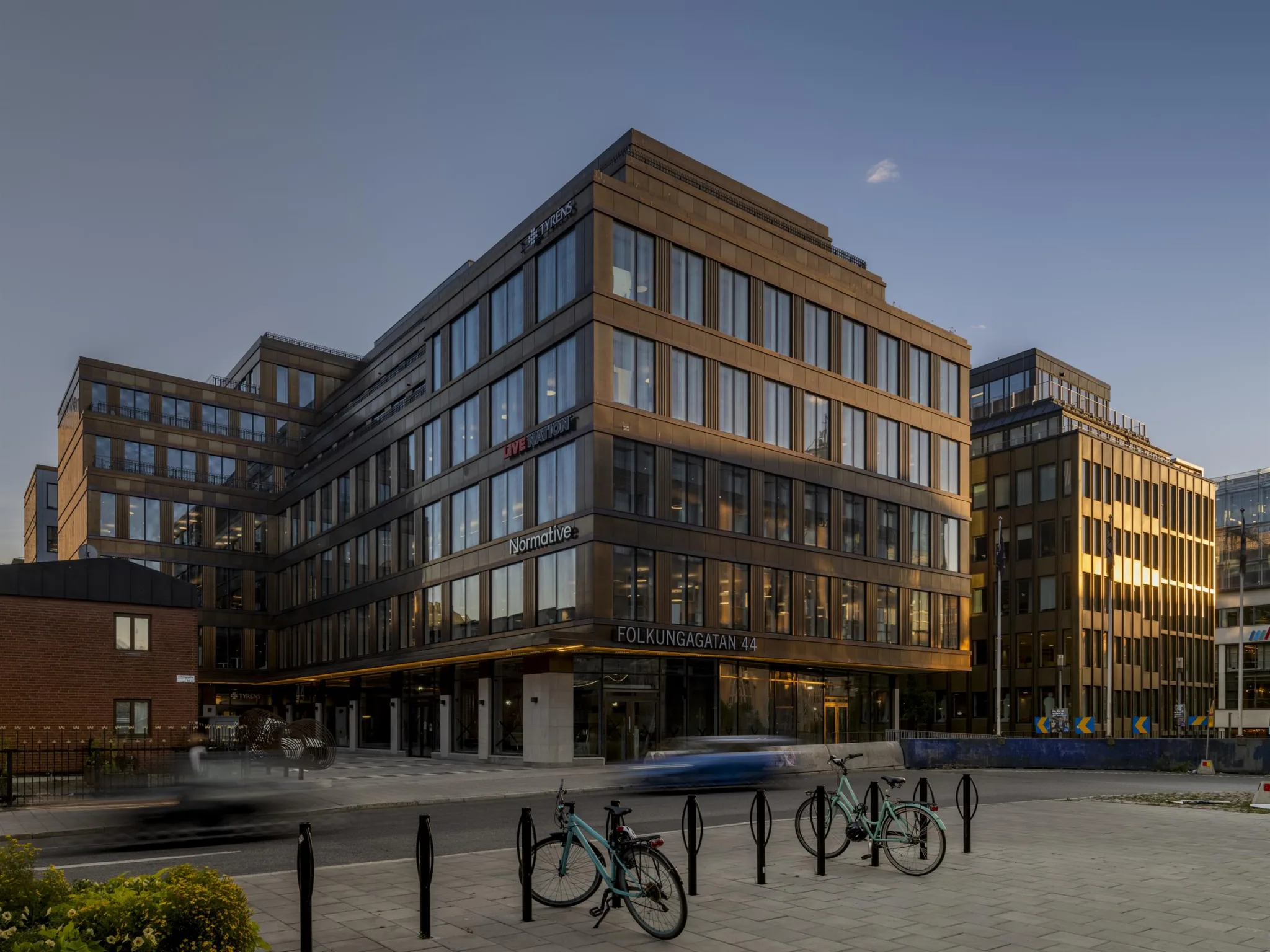 En modern byggnad med stora glasfönster, omgiven av cykelställ och bilar i stadsmiljö.